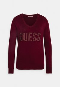 Burgunderfarbener Pullover mit langen Ärmeln und V-Ausschnitt, der das Wort "GUESS" in metallischen Nieten über der Brust trägt. Weiche Strickstruktur.