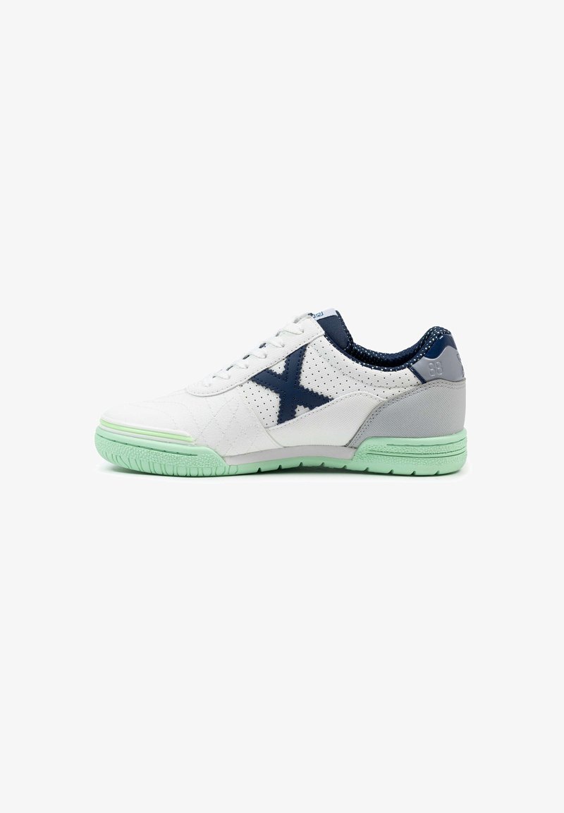 Zapatilla deportiva blanca con textura acolchada, detalles en azul marino, tapa de talón gris y suela de goma verde menta. Presenta secciones perforadas para una mejor transpiración.