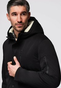 Abrigo negro con capucha que presenta un cuello forrado de forro polar, cremallera frontal y bolsillos laterales con cremallera. La textura es suave con un diseño estructurado.