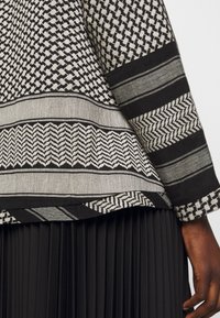 Detailaufnahme eines ärmels und Saums einer schwarz-weiß gemusterten Jacke über einem schwarzen, plissierten Rock, mit einer dunkelhäutigen Hand, die seitlich sichtbar ist.