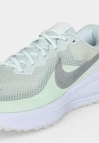 Ανοιχτό γκρι και λευκό αθλητικό παπούτσι Nike με διχτυωτό επάνω μέρος, γκρι λογότυπο Swoosh, λευκά κορδόνια και χοντρή επενδεδυμένη σόλα σε απλό φόντο.