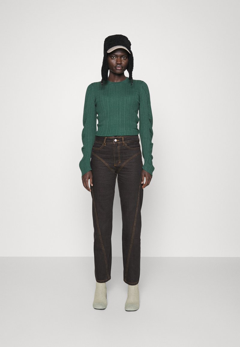 Dunkelbraune Jeans mit sichtbaren Steppnähten, kombiniert mit einem grünen Rippstrickpullover und graugrünen Stiefeletten. Hut mit Ohrenschützern.