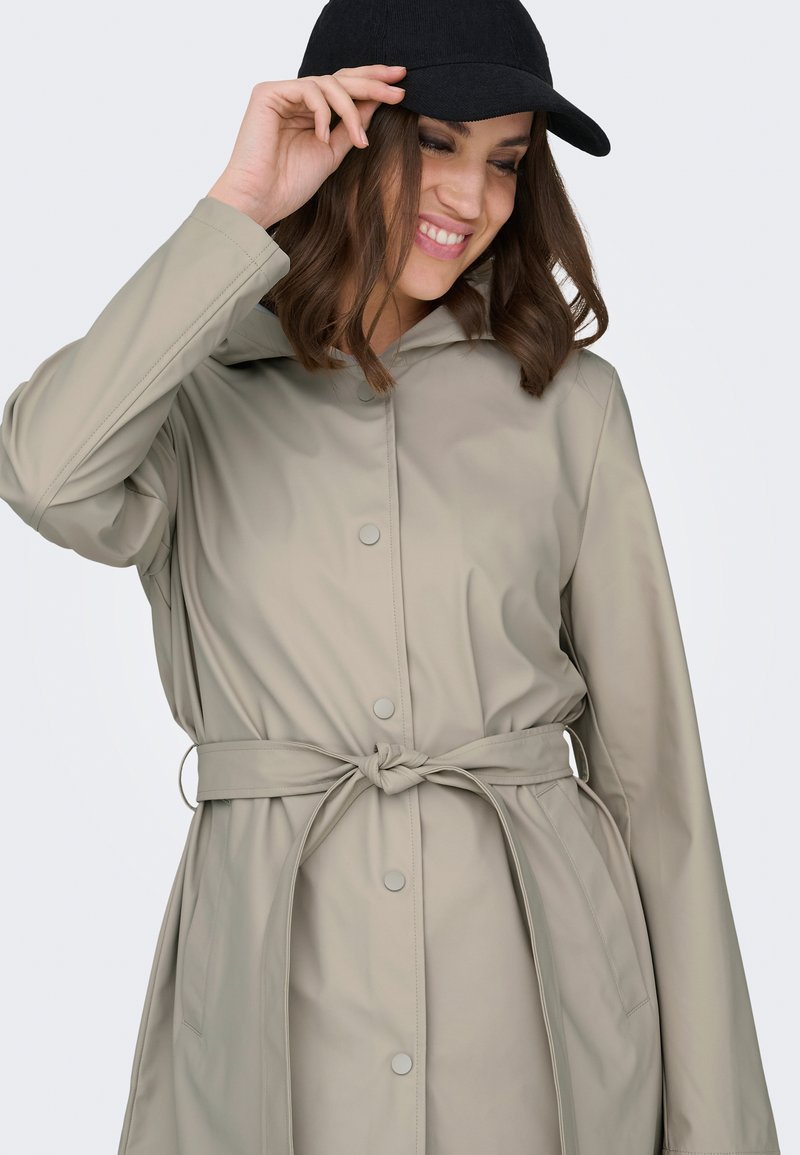 Mujer sonriendo, llevando un impermeable beige con cinturón y ajustando la visera de una gorra de béisbol negra frente a un fondo claro y liso.