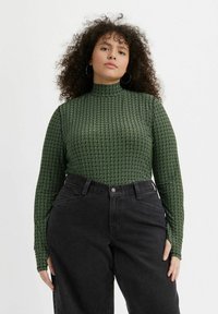 Grøn mønstret langærmet top med høj hals, med små geometriske former; parret med sorte high waist jeans.