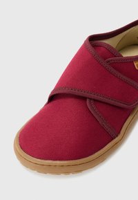 Rød stofsko med rund tå og justerbar Velcro-rem. Har en beige gummisål og tekstureret canvas-overdel for åndbarhed.