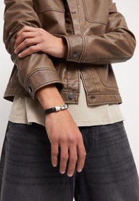 Chaqueta de cuero marrón con puños abotonados, sobre una camisa beige, combinada con jeans gris oscuro. Pulsera negra trenzada con cierre de plata.
