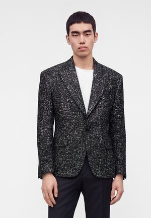 Jeune homme portant un blazer texturé noir et blanc sur une chemise blanche et un pantalon noir, debout devant un fond clair uni.
