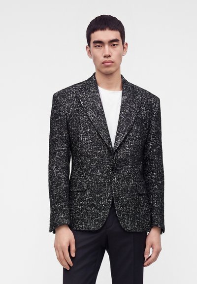 Jeune homme portant un blazer texturé noir et blanc sur une chemise blanche et un pantalon noir, debout devant un fond clair uni.