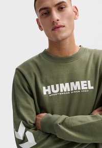 Sudadera verde oliva de mezcla de algodón con el logo y texto blanco "HUMMEL", con distintivos patrones de chevrón blancos en las mangas.