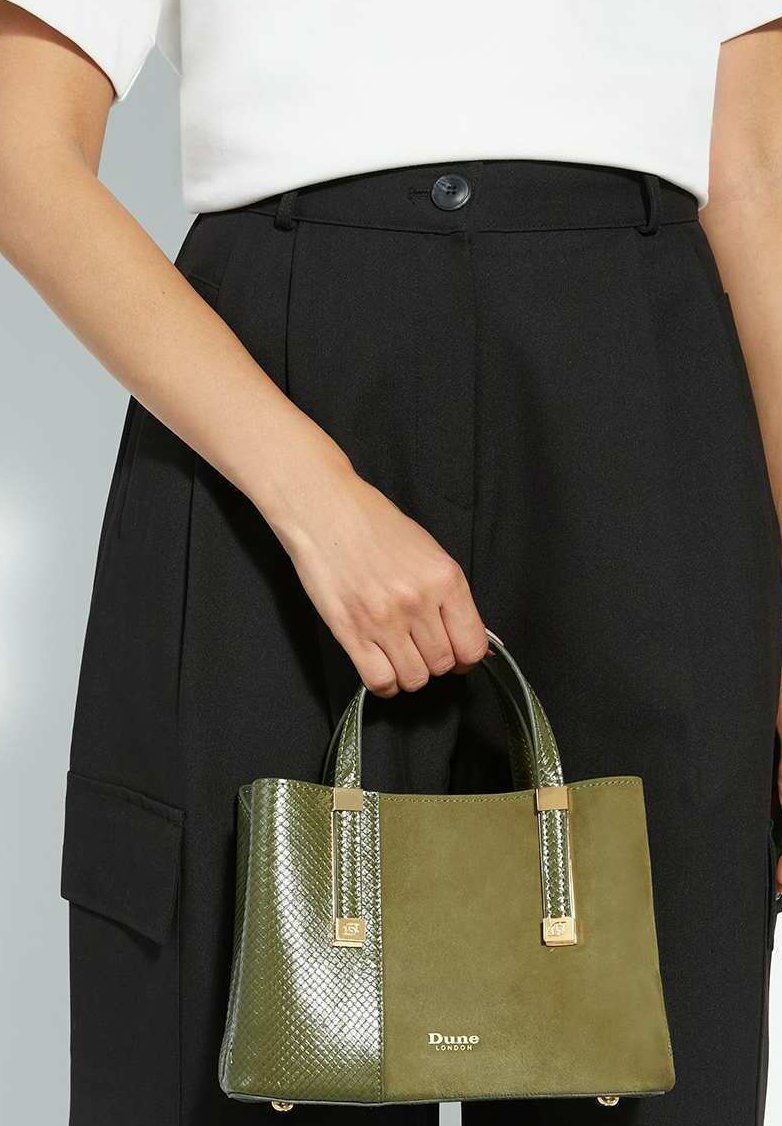 Een hand houdt een kleine bi-kleurige olijfgroene handtas van Dune London vast, met textuur en suède panelen, gecombineerd met zwarte broek en een wit topje.