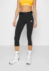 Donna che indossa leggings neri Adidas a tre quarti, reggiseno sportivo arancione, calzini bianchi Adidas e scarpe da corsa bianche, in piedi su uno sfondo bianco.