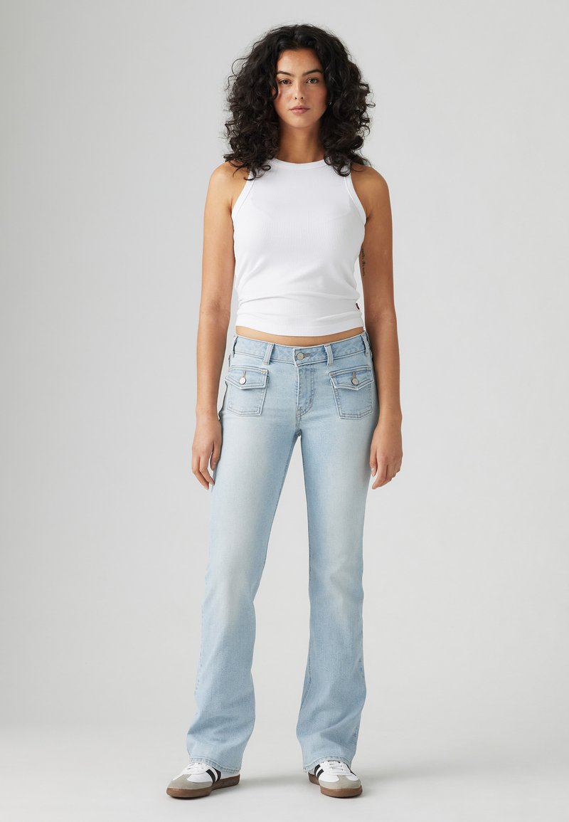 Jean bootcut en denim bleu clair avec deux poches avant, une fermeture à bouton et une finition délavée ; associé à un haut ajusté sans manches blanc.