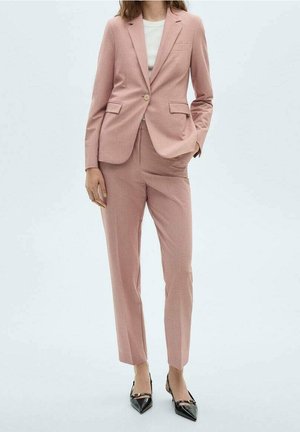 Femme portant un tailleur rose clair sur mesure avec un blazer à un bouton, un haut blanc, un pantalon assorti et des talons noirs pointus.
