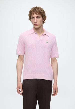 Bărbat cu păr creț blond-închis, purtând un tricou polo roz texturat cu mâneci scurte și pantaloni închiși la culoare, stând în fața unui fundal alb.