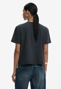 Schwarzes Baumwoll-T-Shirt mit kurzen Ärmeln, lockerem Schnitt und geradem Saum. Getragen mit blauen Jeans mit Gesäßtaschen.