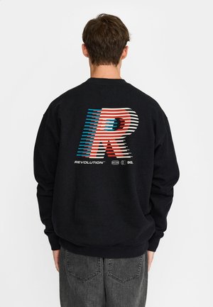 Man draagt een zwart sweatshirt met een grote rood-wit-blauwe gestileerde letter "R" en tekst "REVOLUTION 95" op de rug.