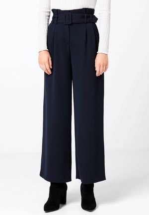 Pantalon taille haute bleu marine à jambes larges avec une ceinture en tissu assortie, porté avec des bottines noires à talons et un haut blanc à manches longues.