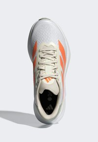 Zapatilla con un upper gris texturizado, rayas naranjas y acentos beige; cuenta con una lengüeta acolchada y una suela con amortiguación.