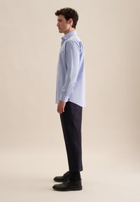 Camicia a maniche lunghe azzurro chiaro con colletto a bottoni, taschino sul petto e polsini; abbinata a pantaloni scuri e scarpe nere. Texture liscia.