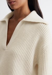 Pull en maille couleur crème avec un large col, un décolleté en V et un motif côtelé texturé. Matière douce et légère avec une coupe décontractée.