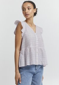 Blouse lilas clair avec des manches courtes, col en V, tissu texturé et boutonnage à l'avant. Présente un ourlet à volants et une coupe douce et fluide.