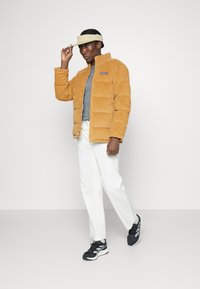 Chaqueta acolchada de pana mostaza con ligero brillo, cremallera frontal, camiseta de cuello redondo gris, pantalones blancos y zapatillas negras con acentos blancos.