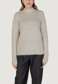 Heller beiger Strickpullover mit hohem Kragen, langen Ärmeln, geripptem Saum und einem strukturierten Muster. Kombiniert mit schwarzen, locker sitzenden Hosen.