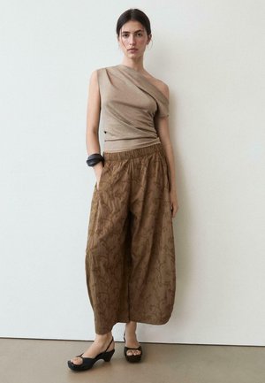 Woman wearing one-shoulder beige top, loose brown floral patterned pants, black open-toe heels, and black chunky bracelet, standing against white wall.