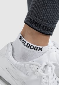 White ankle sock with black "SMILODON" text, paired with gray ribbed leggings and white sneakers, highlighting fabric texture and contrast.