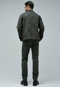 Donkergroene canvasjas en bijpassende slim-fit broek, beide met een gladde textuur en minimalistisch ontwerp. Zwarte laarzen maken de outfit compleet.