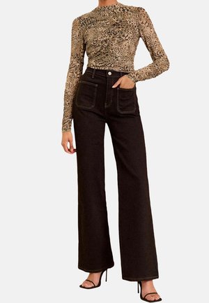 Vrouw die een luipaardprint top met lange mouwen draagt, zwarte wijde broek met hoge taille en voorzakken, en zwarte open teen hakken, staand met één hand in de zak.