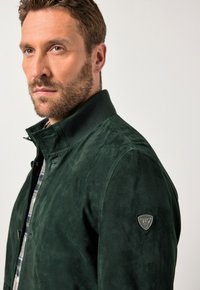 Uomo con capelli corti e barba che indossa un giacca in suede verde scuro con colletto alzato e un piccolo emblema sul braccio superiore.