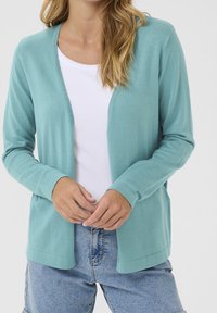 Kobieta ubrana w jasnoturkusowy sweter z otwartym przodem na białą koszulę i jasnoniebieskie dżinsy, ręce delikatnie splecione na wysokości talii.