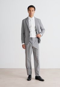 Traje gris claro con americana ajustada y pantalones, camisa blanca debajo. Zapatos negros completan el conjunto. Tejido suave y diseño a medida.