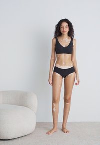 Crni sportski grudnjak i bikini set; glatka tekstura; jednostavan dizajn; model stoji na bež tepihu pored okrugle, svijetle stolice.