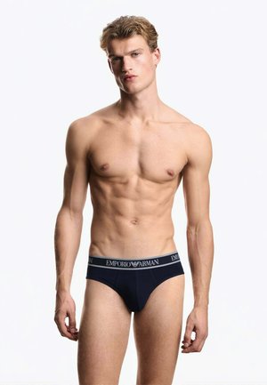 Giovane uomo con corporatura tonica che indossa slip neri Emporio Armani su sfondo bianco semplice.