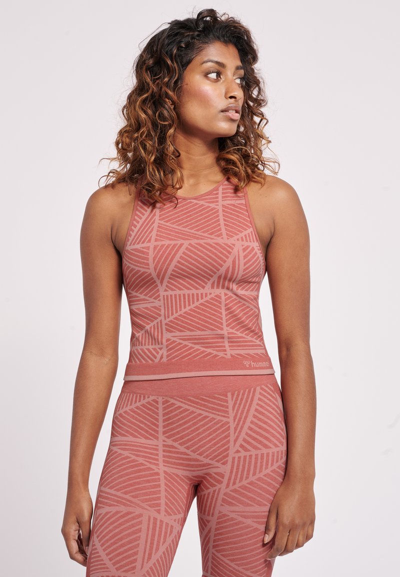 Frau mit lockigem Haar, die ein ärmelloses, gemustertes korallenfarbenes Sporttop und passende Leggings trägt, schaut nach rechts vor einem schlichten Hintergrund.