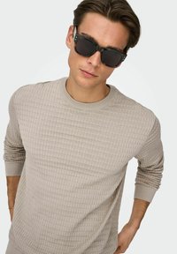 Pull en tricot beige texturé avec poignets et encolure côtelés, associé à des lunettes de soleil noires. Monture carrée avec verres foncés.