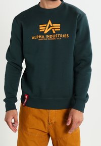 Sudadera verde oscuro con cuello redondo, que presenta un logo naranja de "Alpha Industries" y una pequeña etiqueta roja en el lateral.