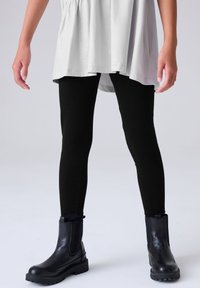 Crne tajice glatke teksture uparene s crnim gležnjačama s debelim potplatom i elastičnim bočnim panelima, nošene ispod svijetlosive tunike.