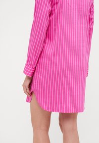 Vestido de camisa a rayas rosas con mangas largas, con dobladillo redondeado, aberturas laterales y rayas blancas verticales. Textura de tela ligera.