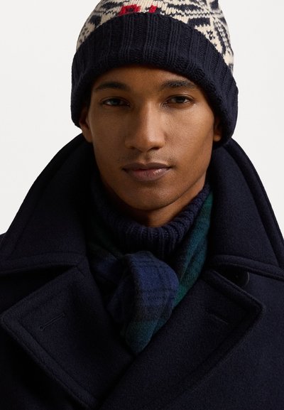 Manteau en laine noire avec de grands revers, bonnet en tricot à motifs bleu marine et écharpe à carreaux vert foncé et bleu, mettant en avant la texture et les superpositions.