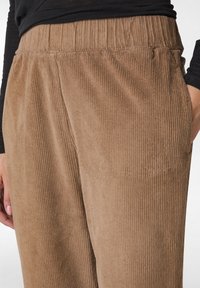 Bruna kordfløyelsbyxor med en ribbad textur, med resår i midjan och sidofickor. Närbild av tyget och passformen vid midjan.