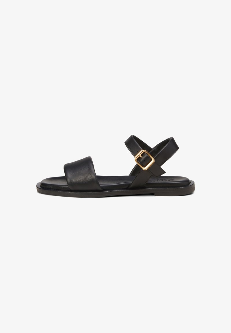 Sandale en cuir noir avec une large bride avant, une bride de cheville ajustable avec une boucle dorée, et une semelle plate. Design simple et minimaliste.
