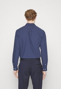 Camicia blu navy con pois bianchi, maniche lunghe, colletto a punta e polsini con bottone. Indossata con trousers scuri, vista posteriore.