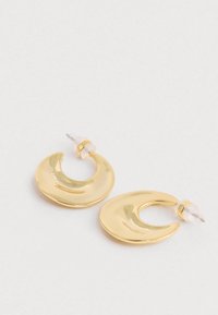 Pár zakrivených, mesiacov tvarovaných leštených zlatých kruhových náušníc s priehľadnými plastovými záchytmi na bielej ploche.