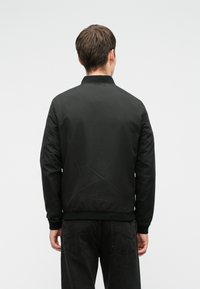 Homme vu de dos portant une veste bomber noire et un jean noir sur un fond gris clair uni.