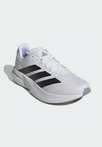 Baskets de sport blanches avec tige en mesh, rayures noires, semelle rembourrée et logo sur le talon. Présente un design à lacets pour un ajustement optimal.