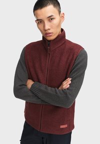 Bordeaux zip-up vest med grå strikkede ærmer. Har høj krave og tekstureret stof, med et lille logo-patch ved kanten.