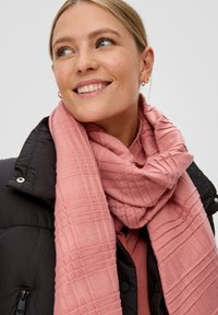 Roze gestructureerde sjaal met ruitpatroon om de nek gewikkeld, gecombineerd met een zwarte pufferjack. Warme, zachte stof met een casual uitstraling.